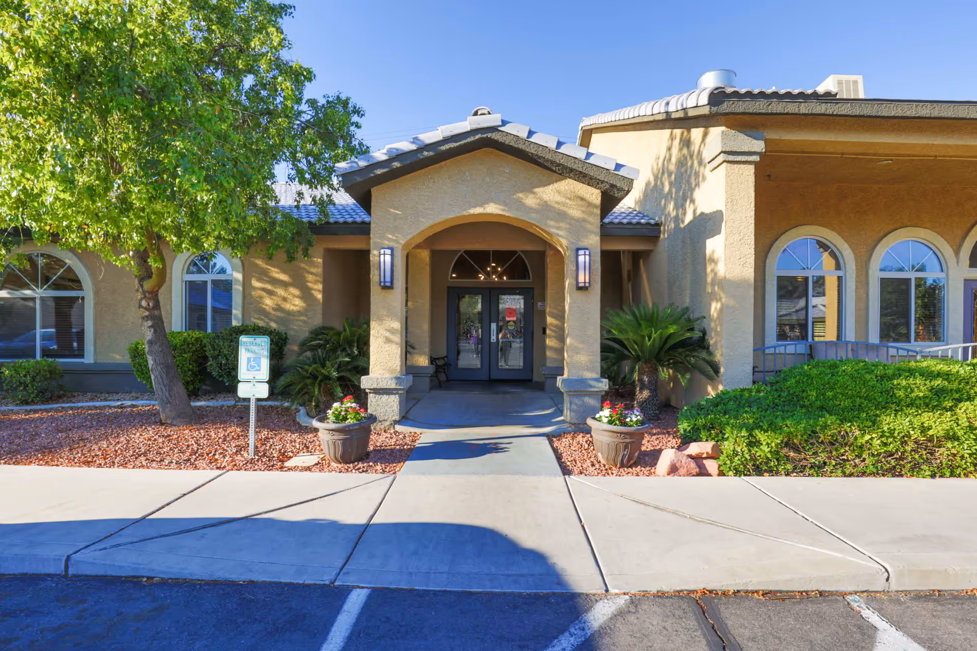 Front entrance of Villa Court Assisted Living facility with a covered walkway, two potted plants with flowers on either side of the entrance, a tree and bushes surrounding the building, and a reserved handicap parking sign visible near the sidewalk.