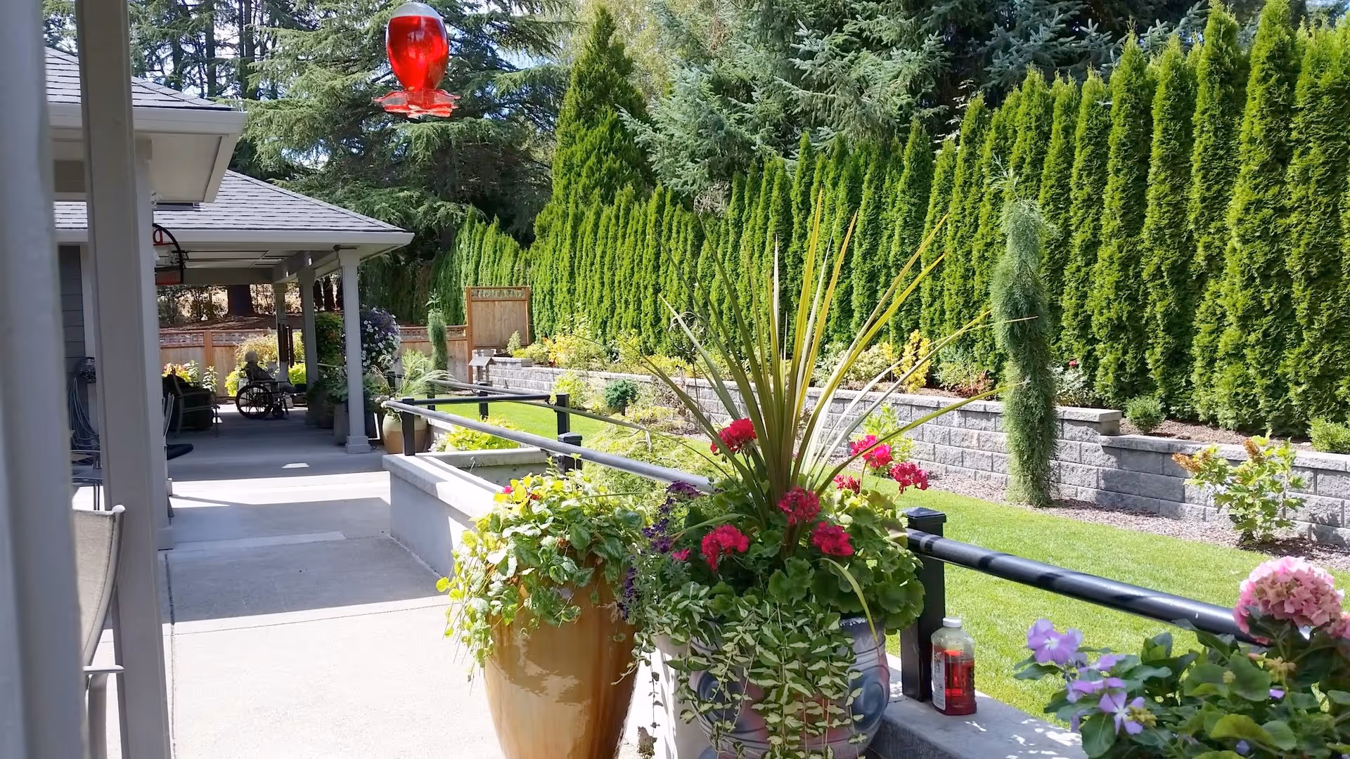 Outdoor patio area at Campan Senior Care Home with potted plants and flowers along a railing, a covered walkway, lush green trees and shrubs in the background, and a red hummingbird feeder hanging above.