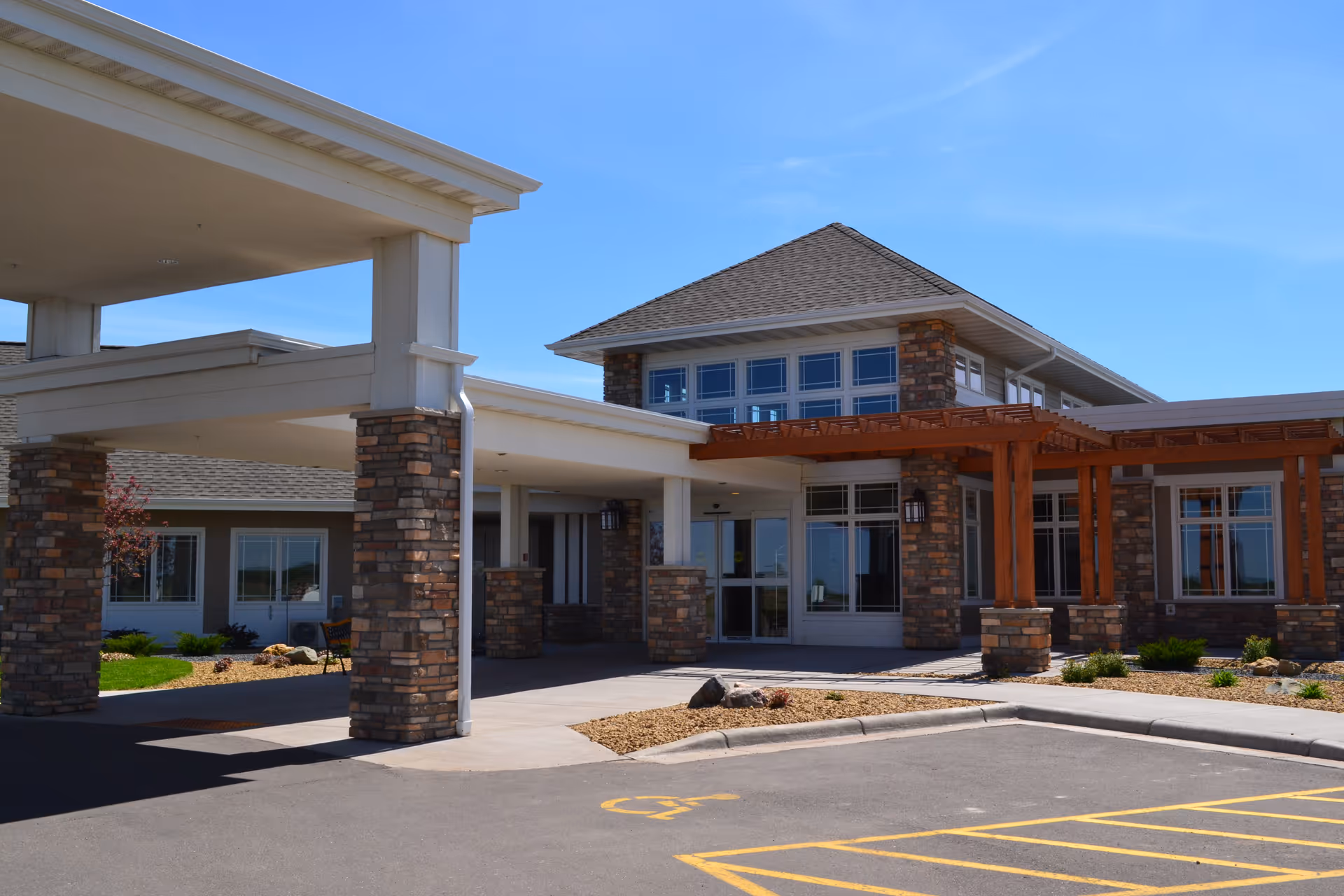 Exterior view of the Christian Community Home of Osceola building featuring a covered entrance with stone pillars, large windows, and a wooden pergola. The foreground shows a parking area with a handicapped parking space marked.