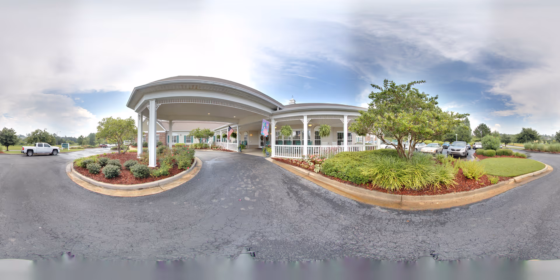 Front entrance of a senior living facility with a covered circular driveway, landscaped islands, and parked cars.