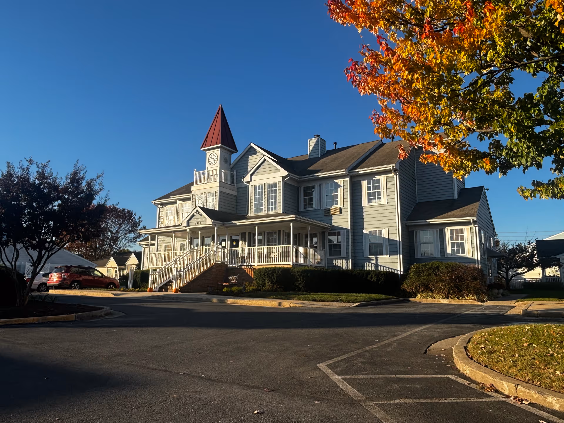 Exterior view of a two-story building with a clock tower and a red pointed roof, surrounded by trees with autumn foliage and a parking area in front under a clear blue sky.