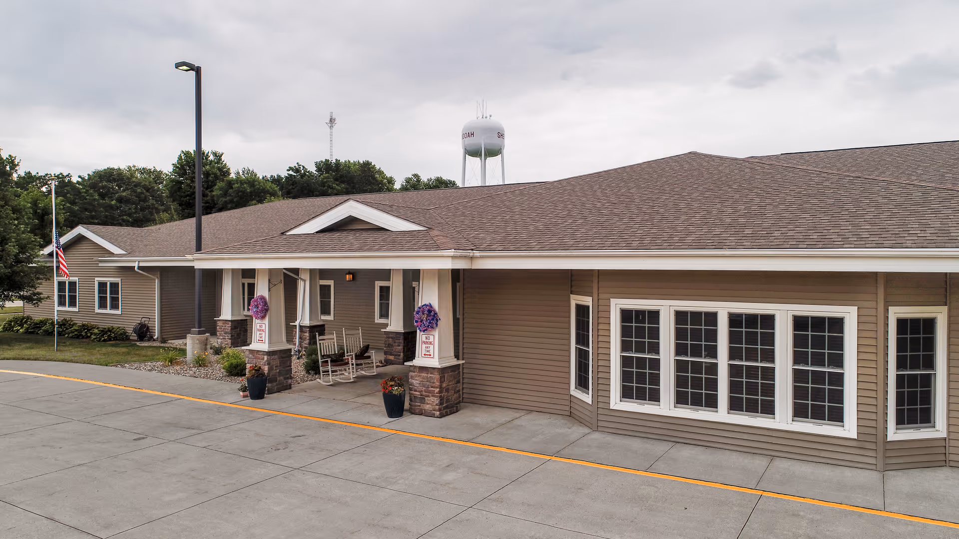 Exterior view of a single-story assisted living facility building with beige siding and a brown shingled roof. The entrance features a covered porch supported by stone pillars with hanging flower baskets and rocking chairs. There is a concrete driveway and an American flag on a pole to the left. Trees and a water tower are visible in the background under a cloudy sky.
