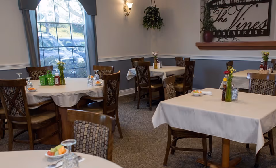 Dining room with multiple tables covered with white tablecloths, each set with glasses, small bowls of fruit, and condiment bottles. Wooden chairs with patterned cushions surround the tables. A large window with blue curtains lets in natural light, and a wall sign reads 'The Vines Restaurant'.