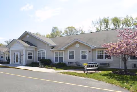 Exterior view of a single-story senior living facility with a bench and blossoming tree.
