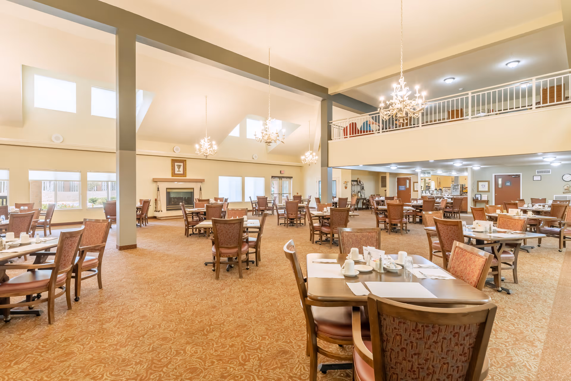 Spacious dining room in a senior living community with multiple wooden tables and chairs arranged neatly. The room features high ceilings with chandeliers, large windows allowing natural light, and a fireplace on one wall. The carpet has a patterned design, and the area appears clean and welcoming.