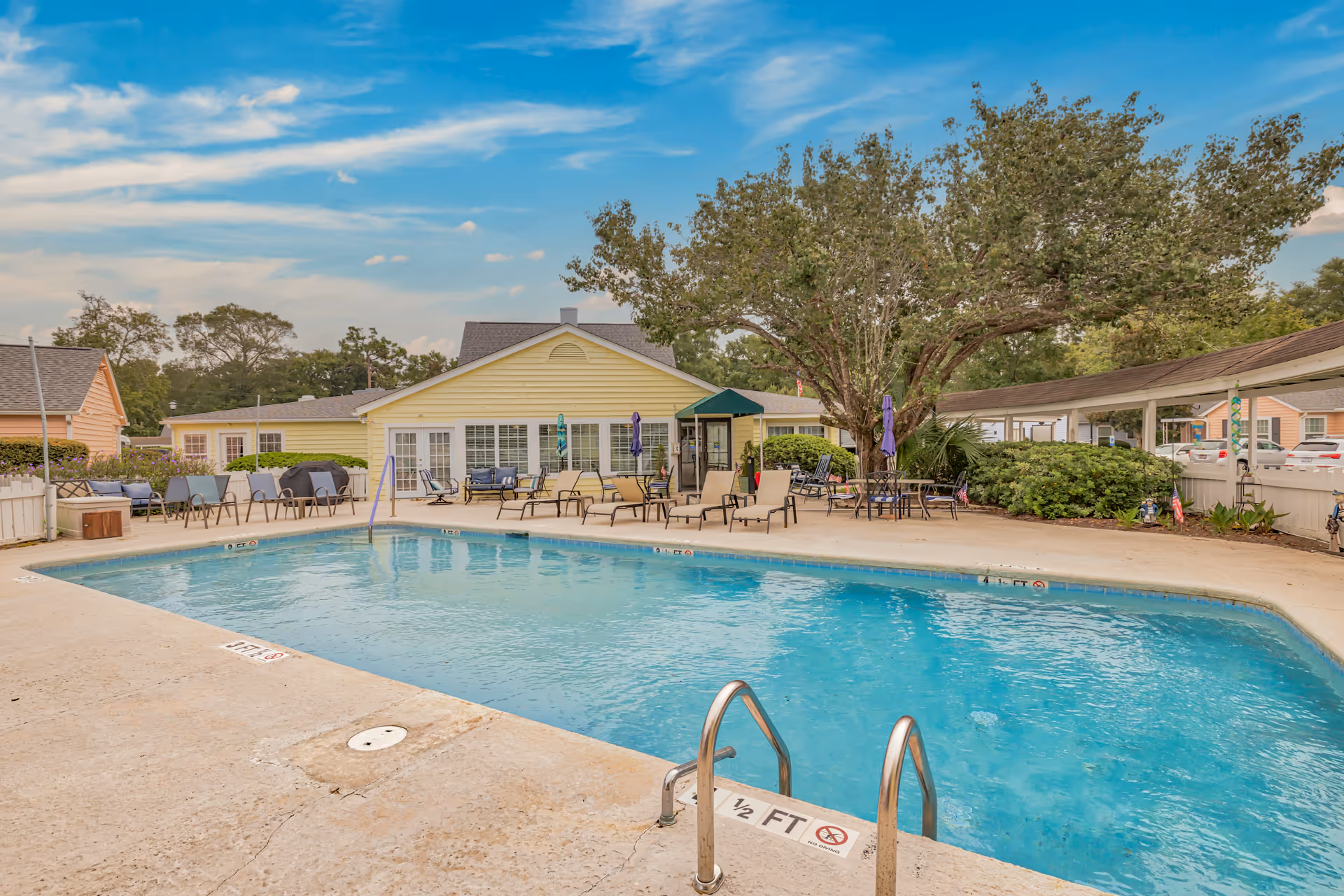 Outdoor swimming pool with clear blue water surrounded by a concrete deck. Several lounge chairs and tables with umbrellas are arranged around the pool. In the background, there is a yellow building with large windows and a green awning, along with trees and parked cars visible behind a white fence.