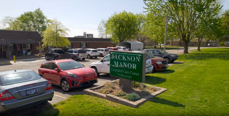 Green 'Jackson Manor' sign in front of a single-story extended care facility, with parked cars and a grassy lawn.