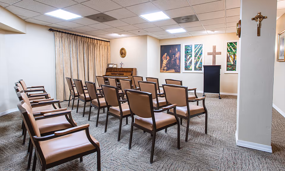 A small chapel or prayer room with rows of brown cushioned chairs facing a piano and a lectern. The room has religious decorations including crosses on the walls and stained glass windows. The floor is carpeted and the ceiling has recessed lighting.