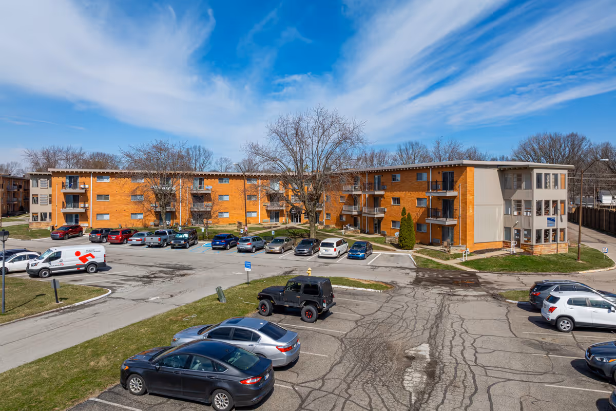 Exterior view of Pinnacle West Apartments, a three-story brick residential building with balconies, surrounded by a parking lot with several parked cars and leafless trees under a partly cloudy blue sky.