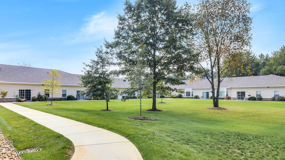 A well-maintained outdoor courtyard area at Maple Lake Assisted Living & Memory Care featuring a curved concrete walkway, green grass, and several trees with a single-story building in the background under a blue sky.