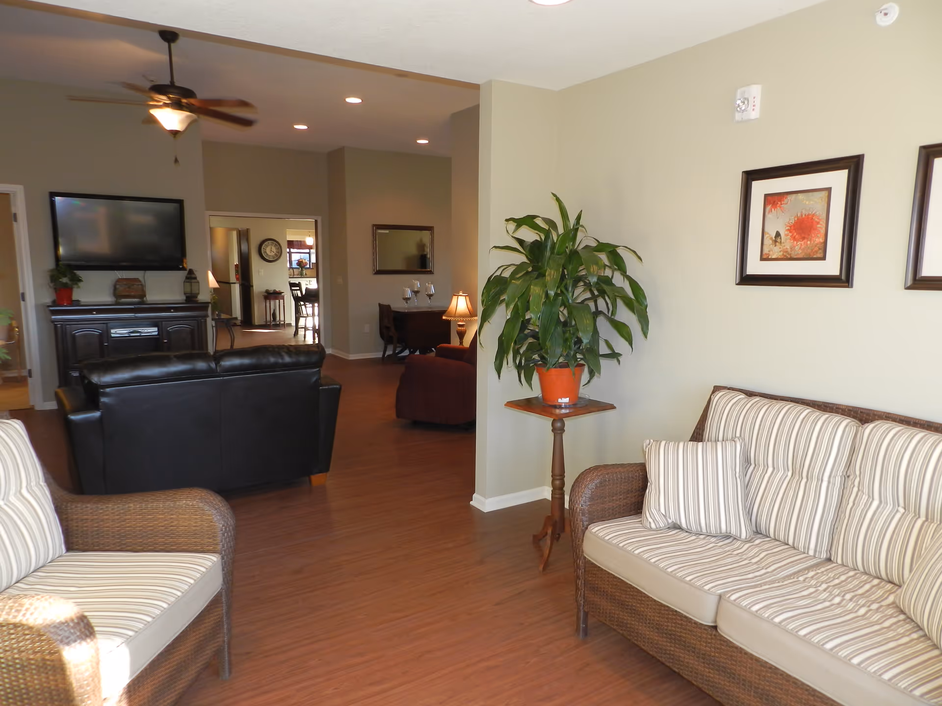 A cozy living room area in Clover Care Home featuring a striped cushioned wicker sofa and chair, a potted plant on a small wooden table, framed artwork on the wall, a ceiling fan, a black leather couch facing a wall-mounted TV, and a view into an adjacent room with more seating and a lamp.