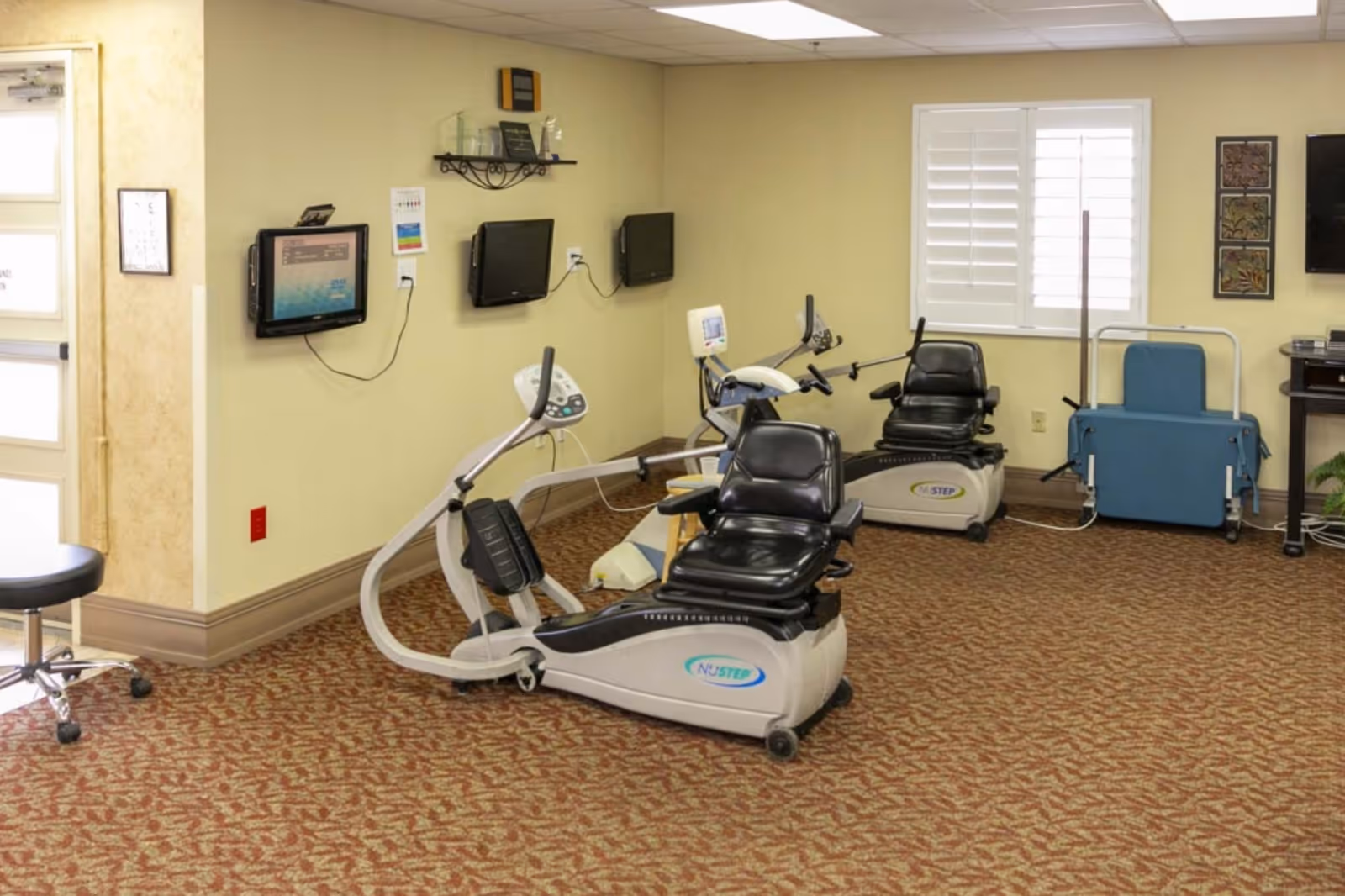 A room with exercise equipment including two NuStep recumbent cross trainers, a blue padded lift chair, and a stool on wheels. The room has beige walls, patterned carpet, a window with white shutters, and three wall-mounted televisions.