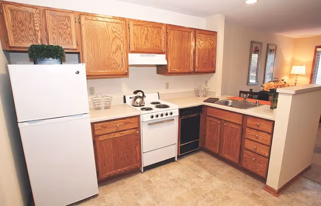 Compact kitchen with oak cabinets, white refrigerator and stove, sink in a corner countertop and view into the adjacent living area.
