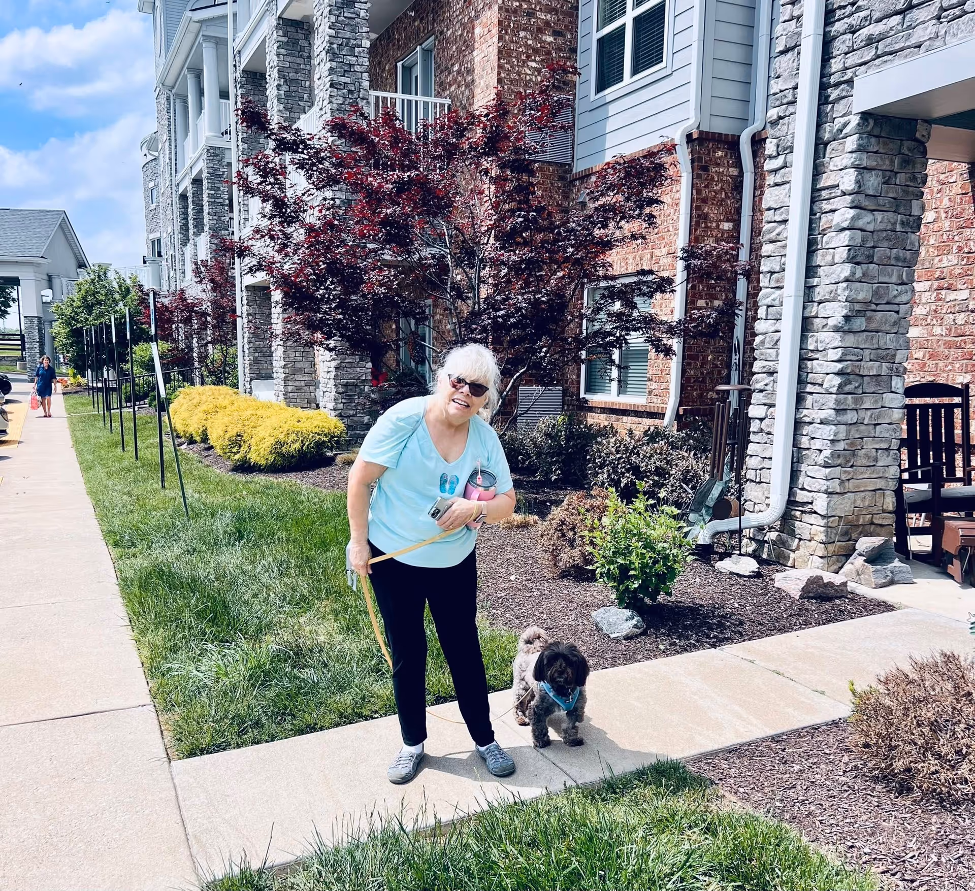An elderly woman with white hair wearing sunglasses, a light blue shirt, black pants, and gray shoes is standing on a sidewalk outside a residential building. She is holding a small dog on a leash and smiling at the camera. The building has a brick and stone exterior with balconies and landscaped bushes and trees. Another person is visible walking in the background along the sidewalk.