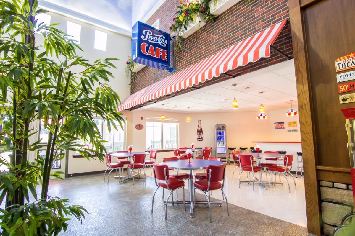 Interior view of a retro-style cafe area with red and white striped awning, red chairs around round tables, a Pepsi-Cola Cafe sign hanging from the ceiling, and a large green plant on the left side. The space has bright lighting and vintage decor including a popcorn machine and wall signs.