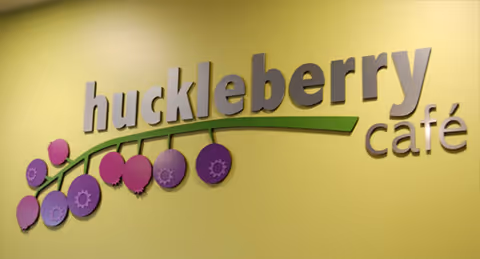 Wall sign with the words 'huckleberry café' in large letters, featuring a green branch with purple and pink circular shapes resembling berries, mounted on a yellow wall.