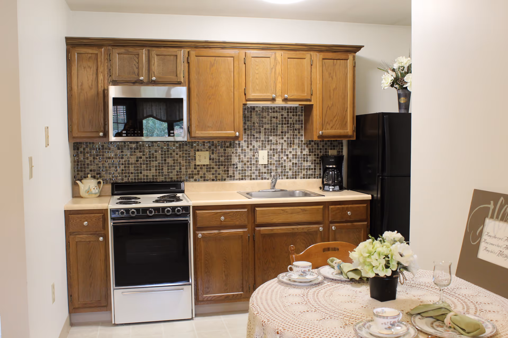 A kitchen area with wooden cabinets, a stove with an oven, a microwave, a sink, a black refrigerator, and a coffee maker. In the foreground, there is a round table covered with a lace tablecloth, set with plates, cups, napkins, and a small flower arrangement.