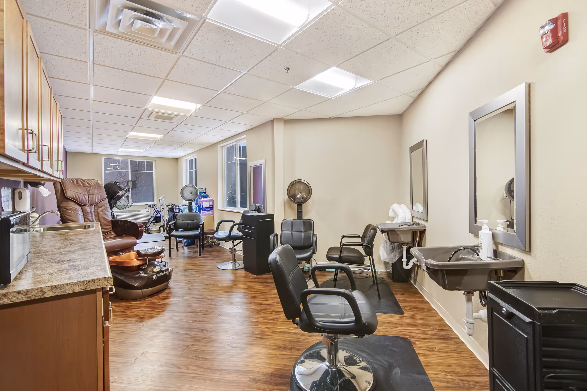 Interior view of a salon area in a senior living facility with multiple black salon chairs, hair drying stations, mirrors, sinks, and wooden cabinets along one wall. The room has wood flooring and large windows letting in natural light.