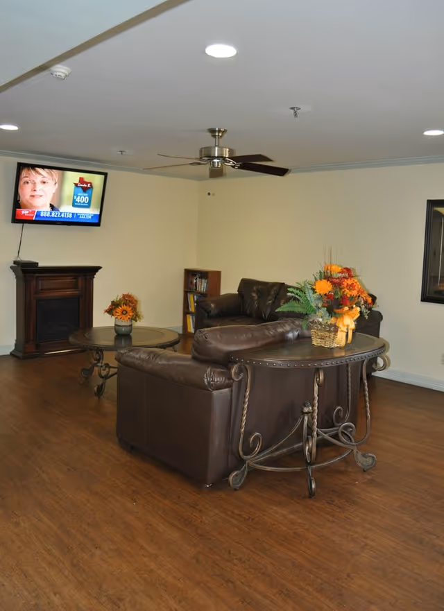 A cozy living room with brown leather sofas arranged around a round coffee table and a side table with a flower arrangement. A flat-screen TV is mounted on the wall above a small fireplace, and a ceiling fan is installed overhead. The room has wooden flooring and light-colored walls.