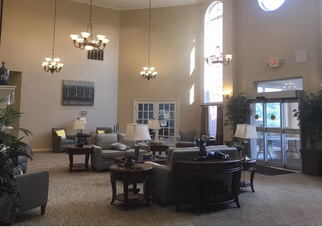 Spacious senior living lounge with high ceilings, chandeliers, sofas and armchairs arranged around side tables near large windows and the main entrance.