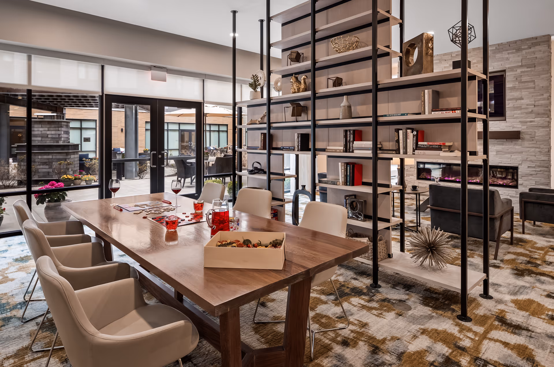 A modern common area with a wooden table surrounded by beige chairs, a game board and drinks on the table, a large open shelving unit with books and decorative items, and a seating area with armchairs and a fireplace in the background. Large glass doors open to an outdoor patio with tables and chairs.