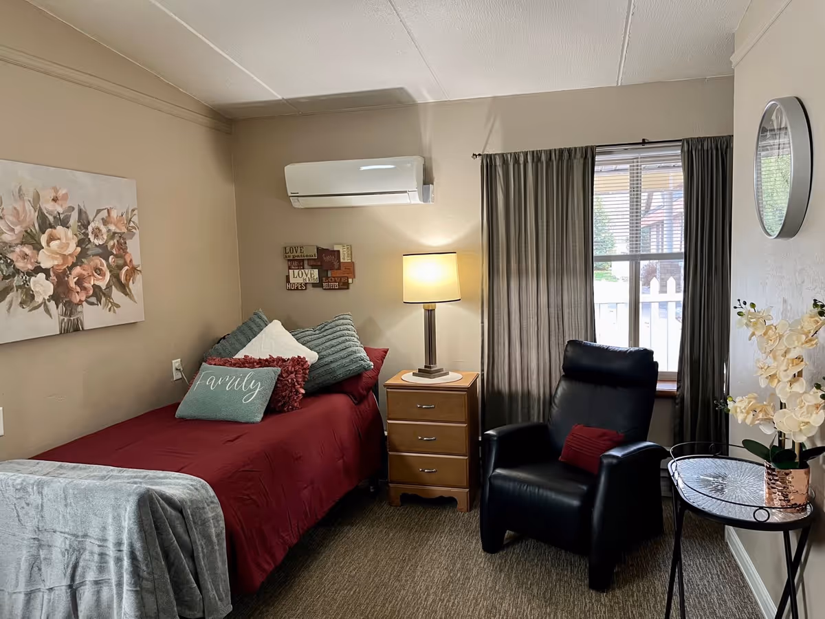 A cozy bedroom with a single bed covered in a red bedspread and multiple pillows, including one with the word 'family'. Next to the bed is a wooden nightstand with a lamp. A black recliner chair is positioned near a window with closed curtains. A small round table with a flower vase is beside the chair. The walls are beige, decorated with a floral painting and a wall clock.