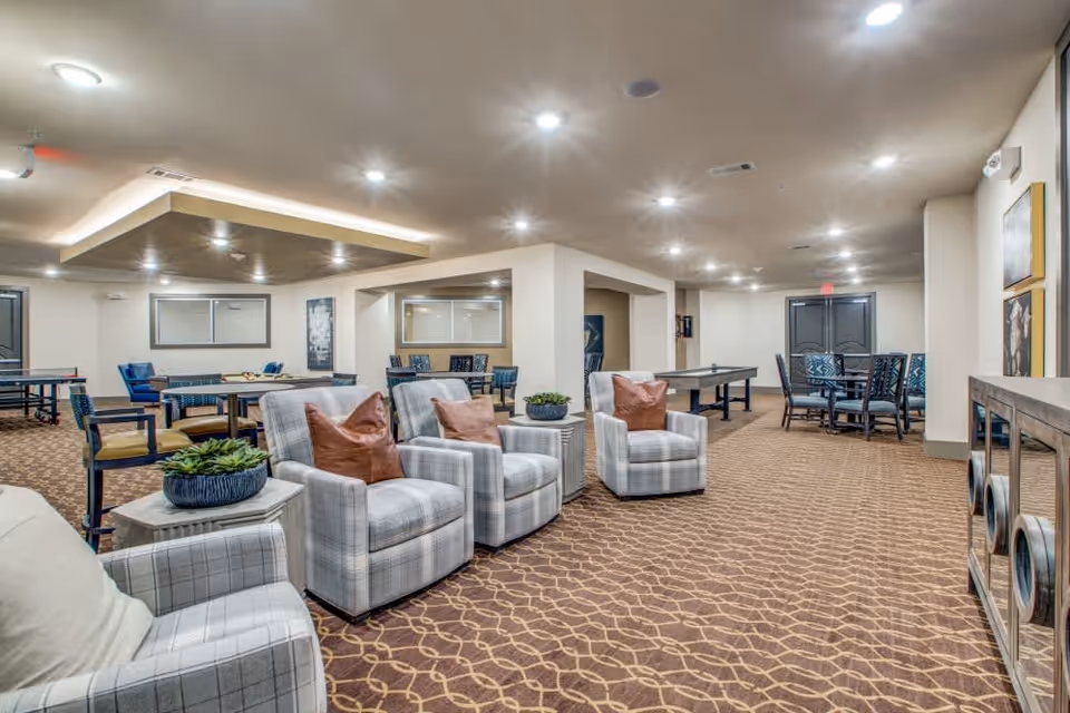 A spacious, well-lit common area with patterned carpet flooring, featuring several plaid armchairs with brown pillows, small side tables with potted plants, multiple tables and chairs for games or socializing, and a ping pong table in the background. The ceiling has recessed lighting and a decorative dropped section with additional lights.