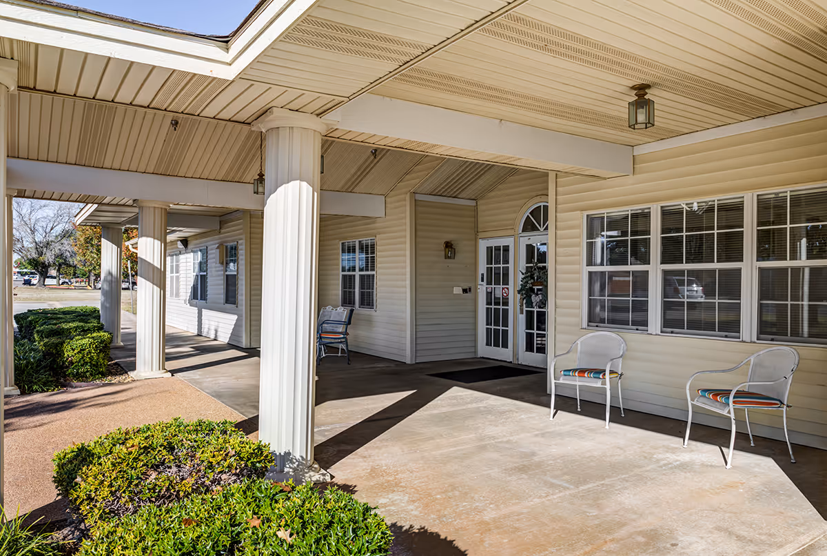 Covered entrance porch of a light-colored building with columns, two chairs, double glass doors, and shrubs.