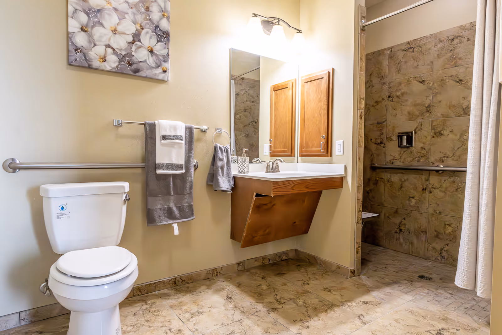 A clean and accessible bathroom featuring a white toilet with a grab bar beside it, a wall-mounted sink with a wooden cabinet underneath, a large mirror above the sink, and a walk-in shower with a curtain and grab bars. The floor and shower walls have a beige marble-like tile pattern. A floral painting hangs on the wall above the towel rack holding gray and white towels.