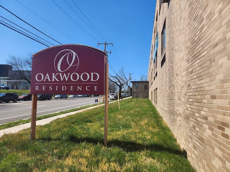Outdoor view of Oakwood Residence sign on a grassy area next to a brick building, with a street and cars in the background under a clear blue sky.