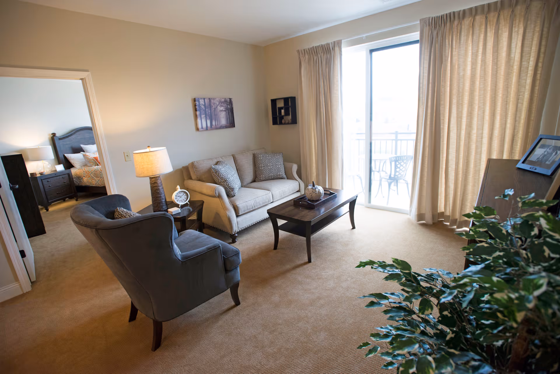 A cozy living room with a beige sofa, two patterned cushions, a dark gray armchair, a wooden coffee table, and a side table with a lamp and clock. The room has beige carpet and light-colored walls. A sliding glass door with beige curtains leads to a balcony with outdoor chairs. A bedroom with a bed and nightstand is visible through an open doorway. A green leafy plant is in the foreground.