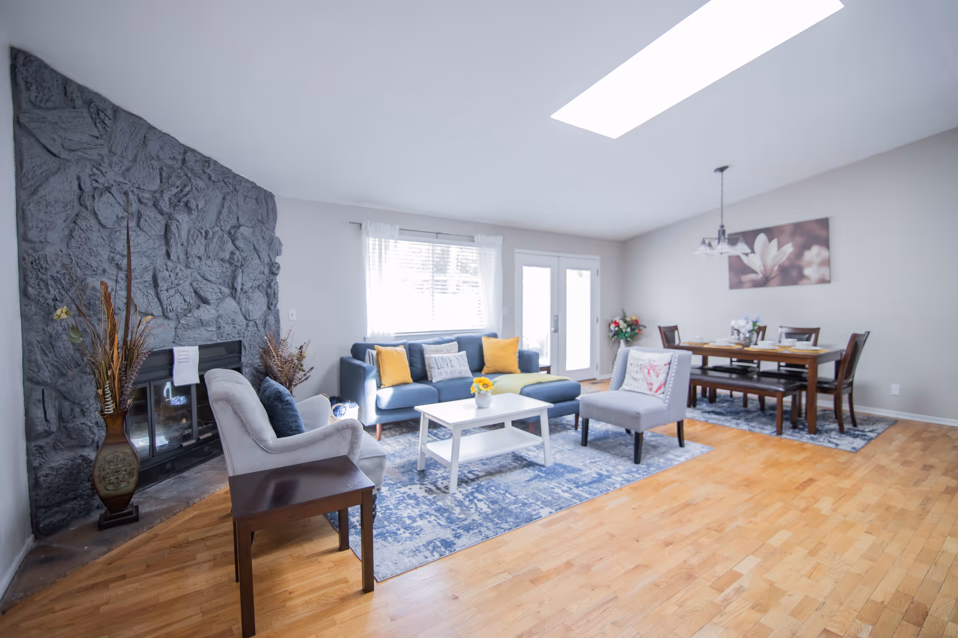 A bright and spacious living and dining area with a stone fireplace on the left, a blue sofa with yellow and white cushions, a white coffee table, and two armchairs. The dining area features a wooden table with chairs and a bench, set with plates and bowls. A large window and glass doors allow natural light to fill the room, complemented by a skylight overhead. The floor is wooden with area rugs under the seating and dining areas, and a floral painting hangs on the wall above the dining table.