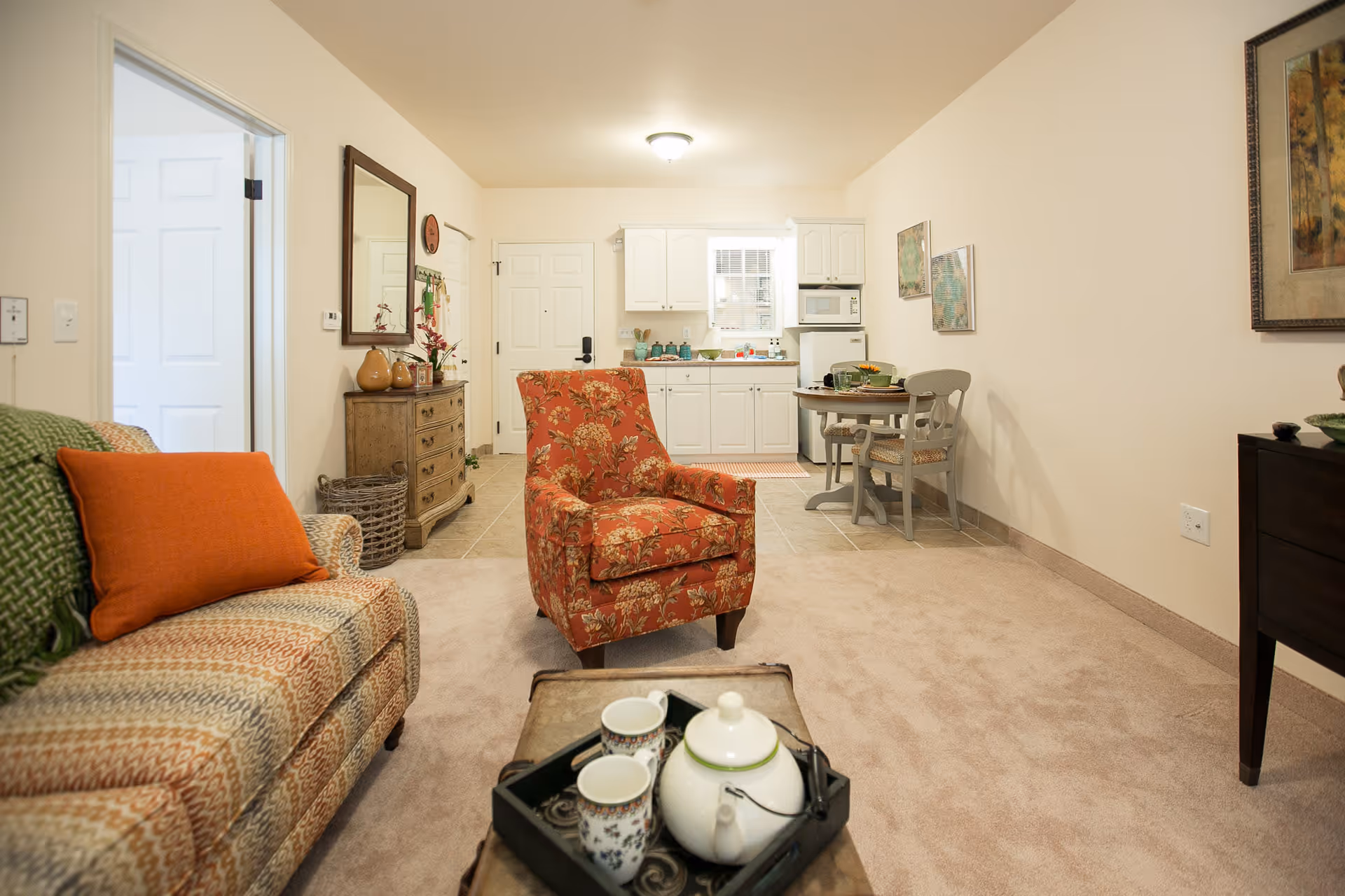 A cozy living area in a senior living facility featuring a patterned sofa with an orange pillow and green throw, a floral armchair, and a wooden coffee table with a teapot and cups. In the background, there is a small kitchen with white cabinets, a microwave, and a round dining table with two chairs. The room has beige walls and carpeted flooring, with a dresser and mirror on one side and framed artwork on the walls.