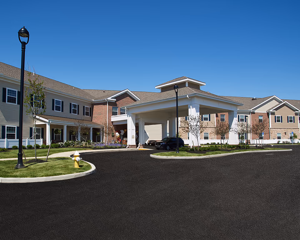 Exterior view of Grove City Senior Living facility showing a large two-story building with beige and brick siding, a covered entrance with white columns, a black car parked under the entrance, street lamps, small trees, and a clear blue sky.