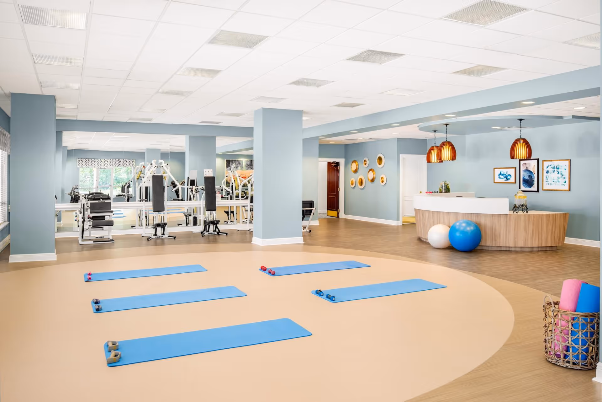 A spacious fitness and wellness room with blue exercise mats and small dumbbells arranged on a beige circular floor area. In the background, there is gym equipment against a mirrored wall. To the right, a reception desk with two large exercise balls and pendant lights hangs from the ceiling. The walls are painted light blue and decorated with framed artwork and circular wall hangings. A basket with rolled yoga mats is visible in the foreground.