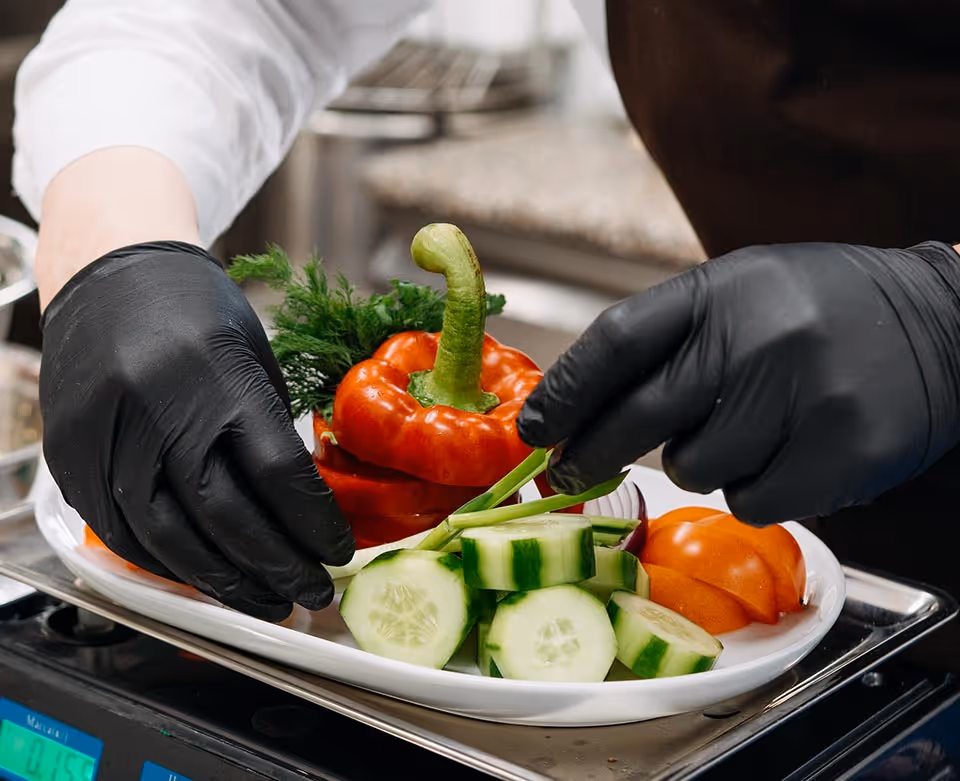 Gloved hands place sliced cucumber and a red bell pepper on a plate in a kitchen.