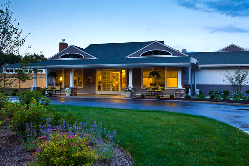 Front entrance of a single-story senior living facility with a covered porch, rocking chairs, and landscaped lawn at dusk.