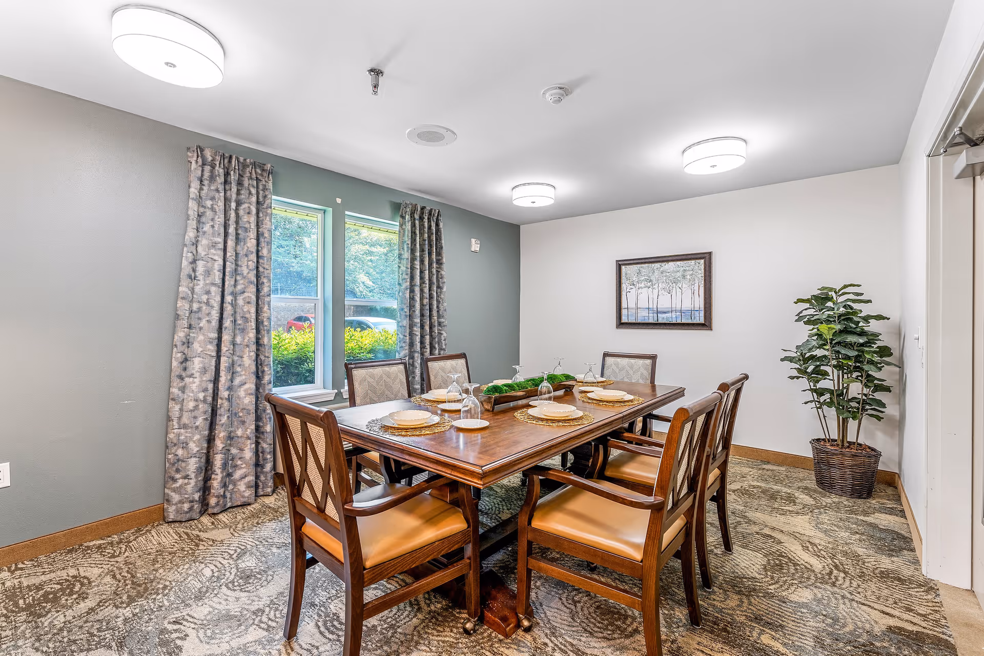 A dining room with a wooden table set for six people with plates, glasses, and placemats. The room has two windows with patterned curtains, a framed picture on the wall, a potted plant in the corner, and ceiling lights providing illumination.