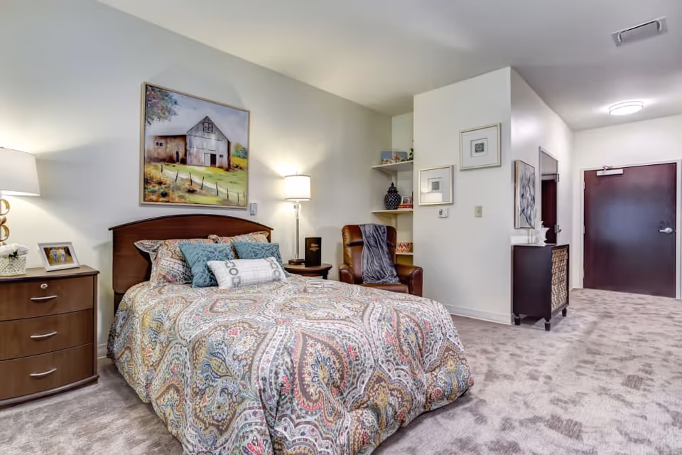 A cozy bedroom in a senior living facility featuring a bed with a colorful paisley-patterned comforter and multiple pillows. There is a wooden nightstand with a lamp and a framed photo on the left side of the bed. A brown leather armchair with a gray throw blanket is positioned near the corner. The walls are decorated with framed artwork, including a painting of a barn above the bed. The room has beige carpeting and a dark wooden door at the far end.