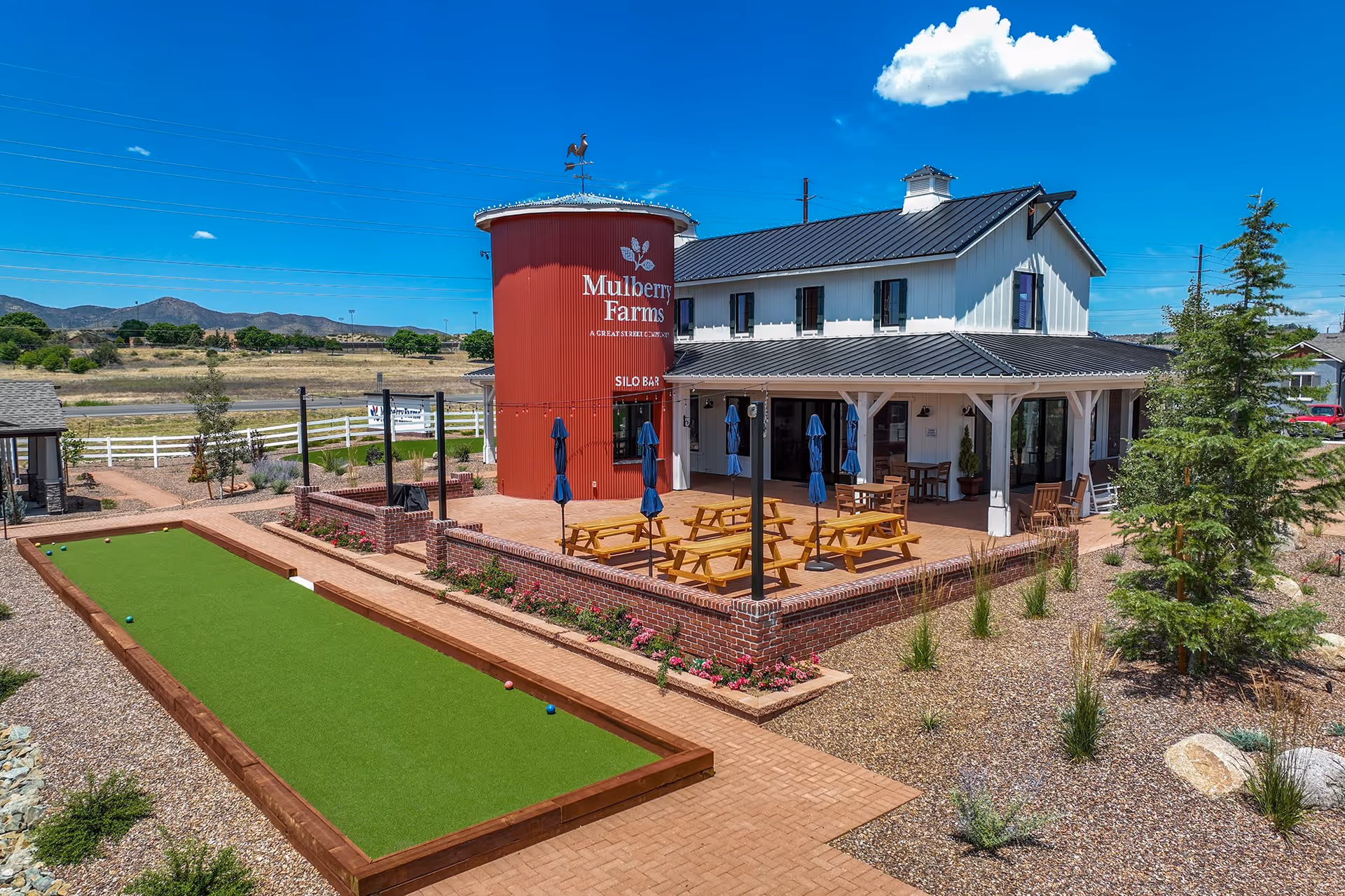 Exterior view of Mulberry Farms showing a red silo, a white two-story building with a covered patio and picnic tables, and a bocce/lawn court in the foreground.