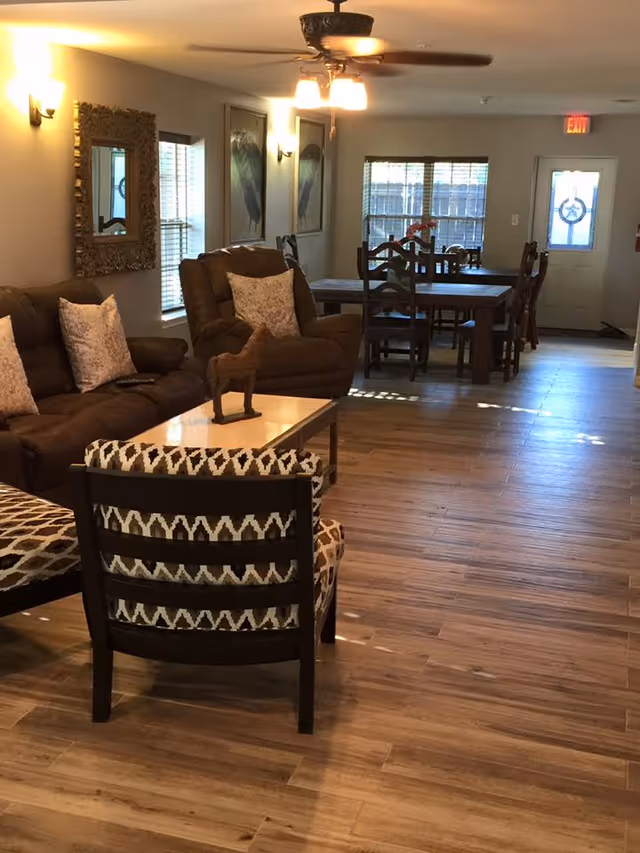 A cozy living and dining area with wooden flooring, featuring a brown sofa with patterned cushions, a matching armchair, a coffee table with a small horse figurine, and a dining table with chairs near windows and a door with a wreath.