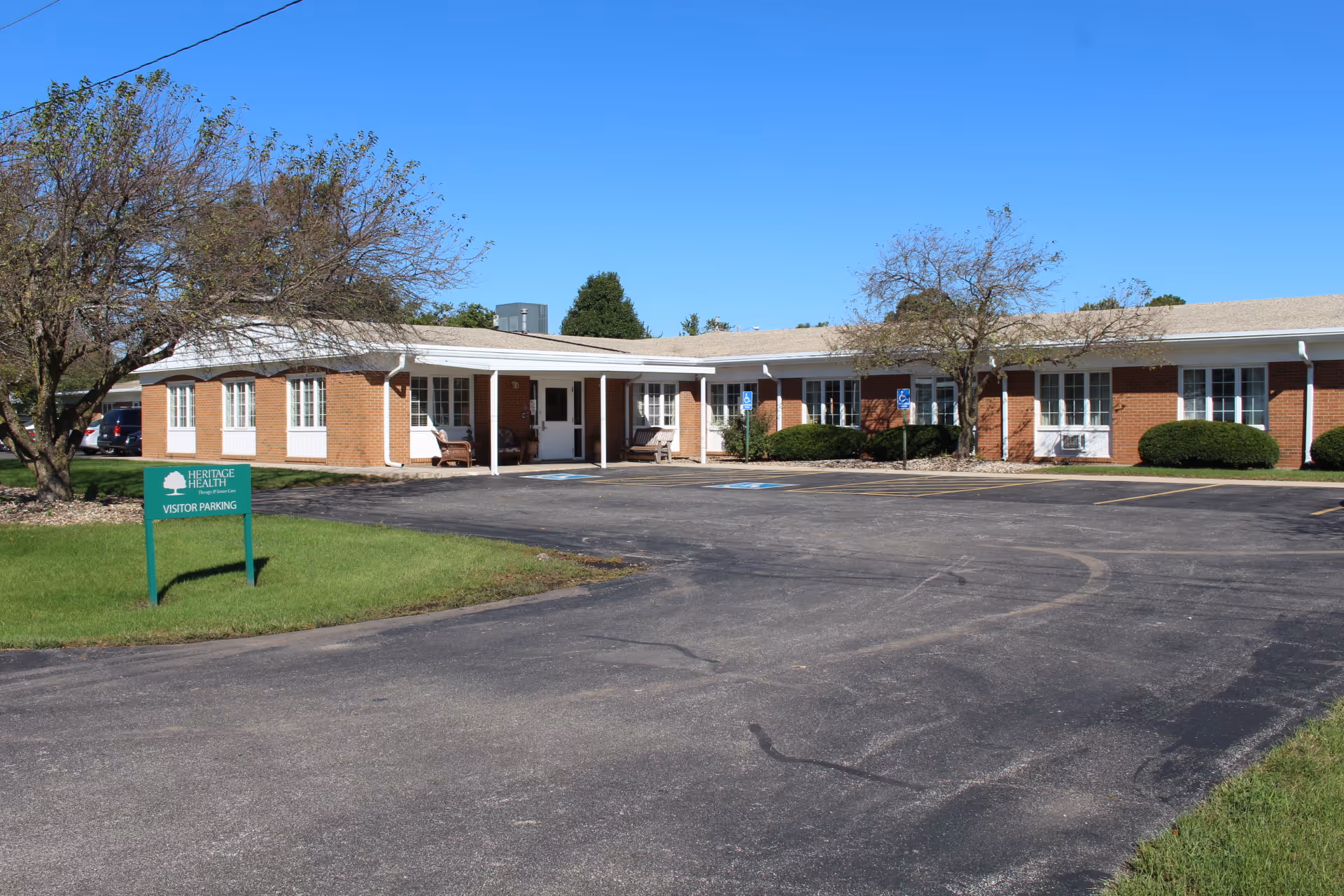 Exterior view of a single-story brick building with white trim and multiple windows, surrounded by trees and bushes under a clear blue sky. There is a paved parking lot in front with marked parking spaces and a green sign that reads 'Heritage Health Visitor Parking.'