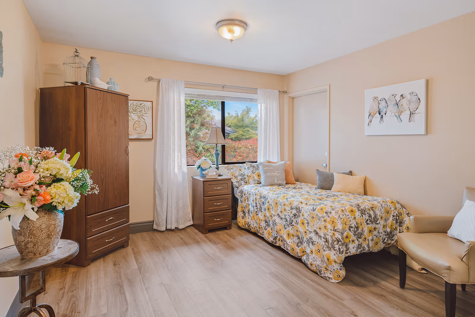 A cozy bedroom with a single bed covered in a yellow and gray floral bedspread, several decorative pillows, a wooden nightstand with a lamp and flowers, a wooden wardrobe, a beige armchair, and a window with white curtains showing greenery outside. The walls are light beige with framed artwork, and the floor is light wood.