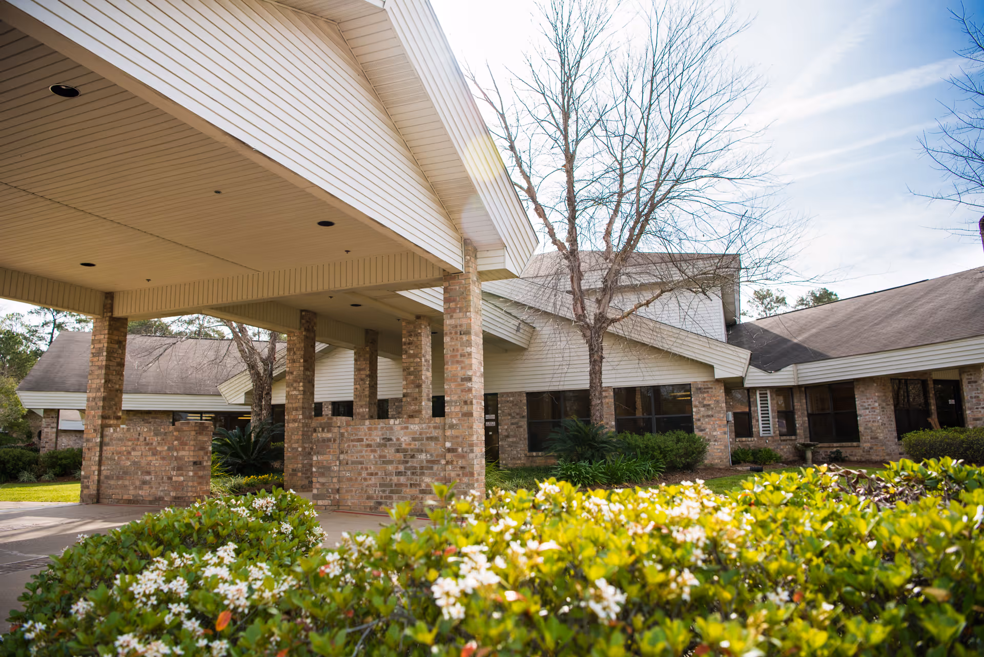 Exterior view of a senior living facility named The Oaks Center showing a covered entrance with brick pillars, leafless trees, and green bushes with white flowers in the foreground under a partly cloudy sky.