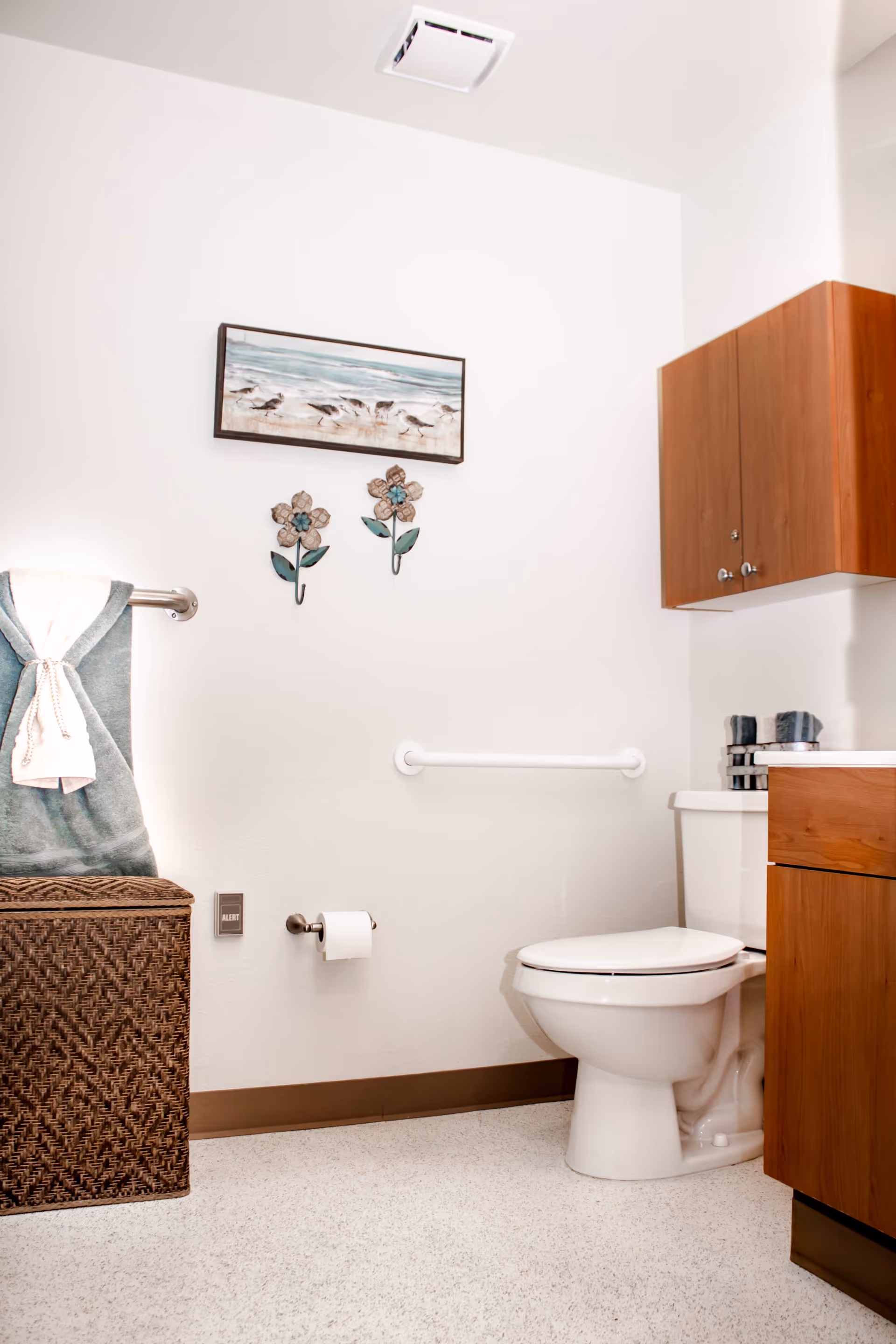 A clean and well-lit bathroom with a white toilet, wooden cabinets, a towel rack holding a folded towel, a woven basket, and wall decorations including a framed picture of birds on a beach and two flower-shaped hooks.