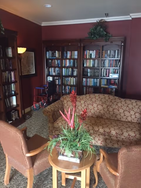 A cozy living room with a patterned sofa and two brown armchairs around a small wooden table with a green and red floral arrangement. Behind the sofa, there are large wooden bookshelves filled with books against a maroon wall. A walker is visible near the bookshelves.