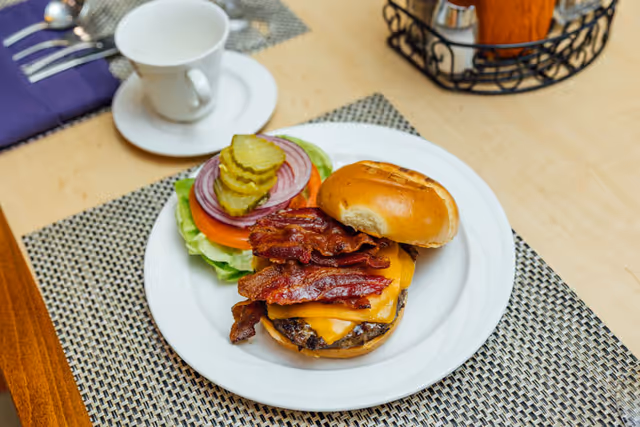 A bacon cheeseburger on a white plate with the top bun slightly lifted, showing crispy bacon, melted cheddar cheese, and a beef patty. On the side of the plate are slices of pickles, red onion, tomato, and lettuce. The plate is on a woven placemat on a wooden table, with a white cup and saucer and a napkin with silverware in the background.