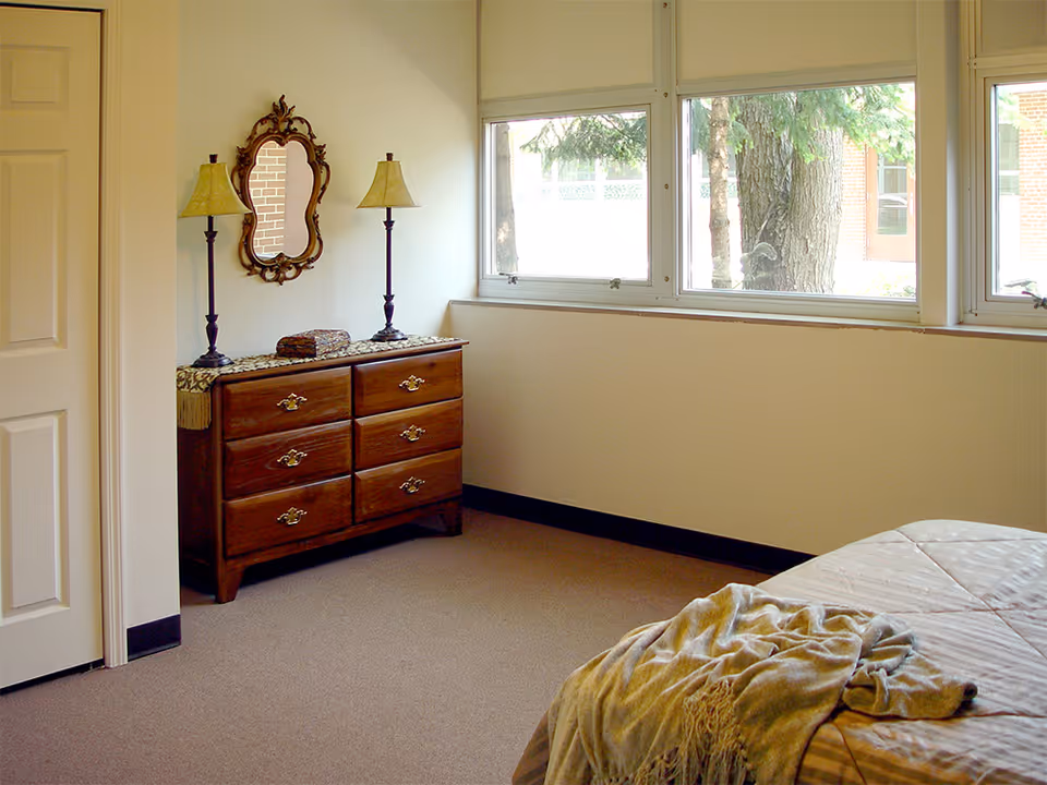 A bedroom corner with a wooden dresser featuring six drawers, two table lamps with beige shades, and an ornate wall mirror above the dresser. There is a large window showing trees and a brick building outside. Part of a bed with a beige blanket is visible in the foreground.