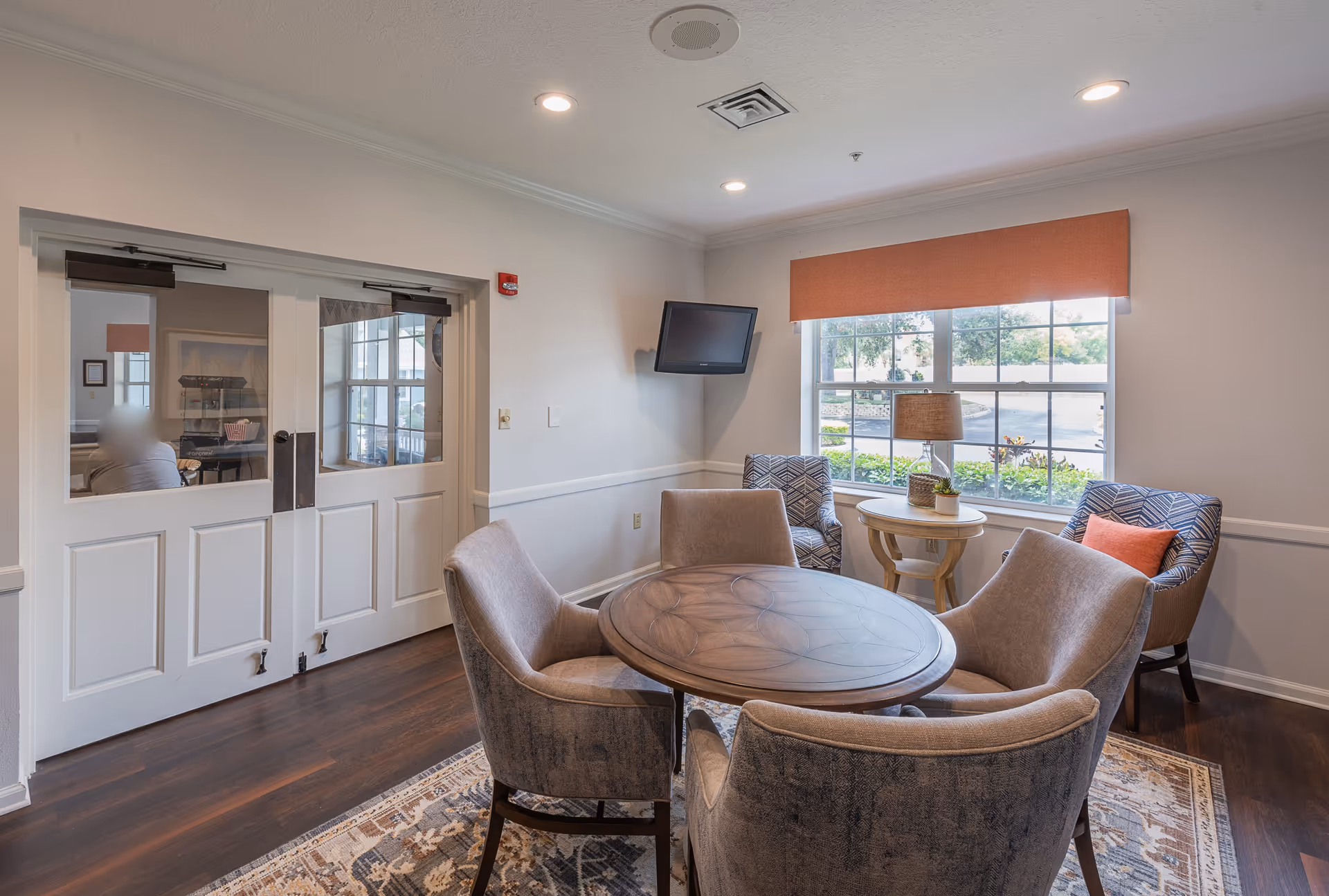 A cozy sitting area in a senior living facility with a round wooden table surrounded by four upholstered chairs. There are two additional patterned armchairs near a window with an orange valance, a small round side table with a lamp and plant, and a wall-mounted TV. The room has wooden flooring and white walls with a double door leading to another room.