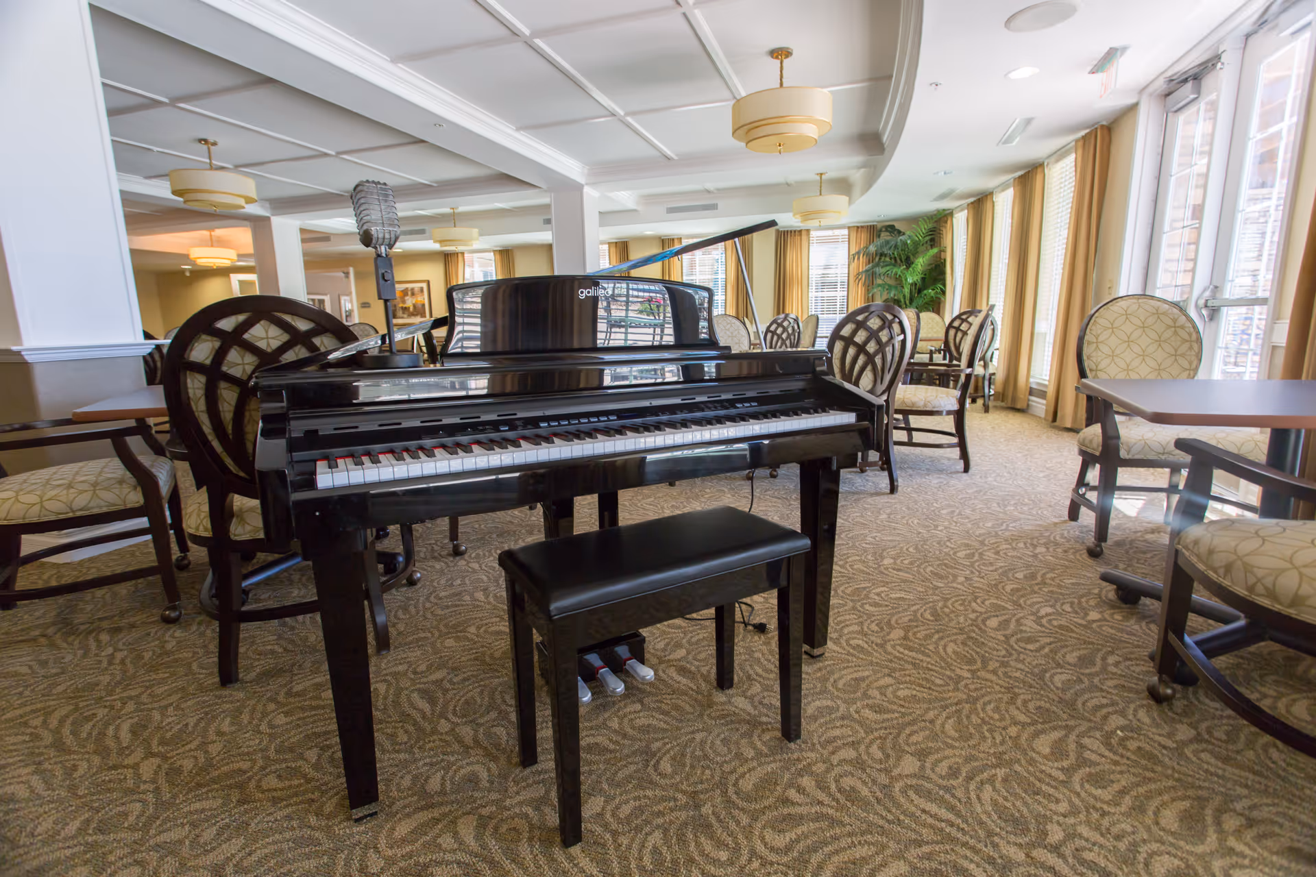 Grand piano and bench in a bright senior living lounge with tables and upholstered chairs by large windows.
